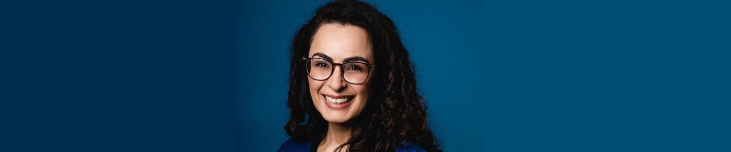 Een vrouw met een bril glimlachend voor een blauwe achtergrond genaamd Amina Saydali wordt de nieuwe directeur/bestuurder. School en Veiligheid