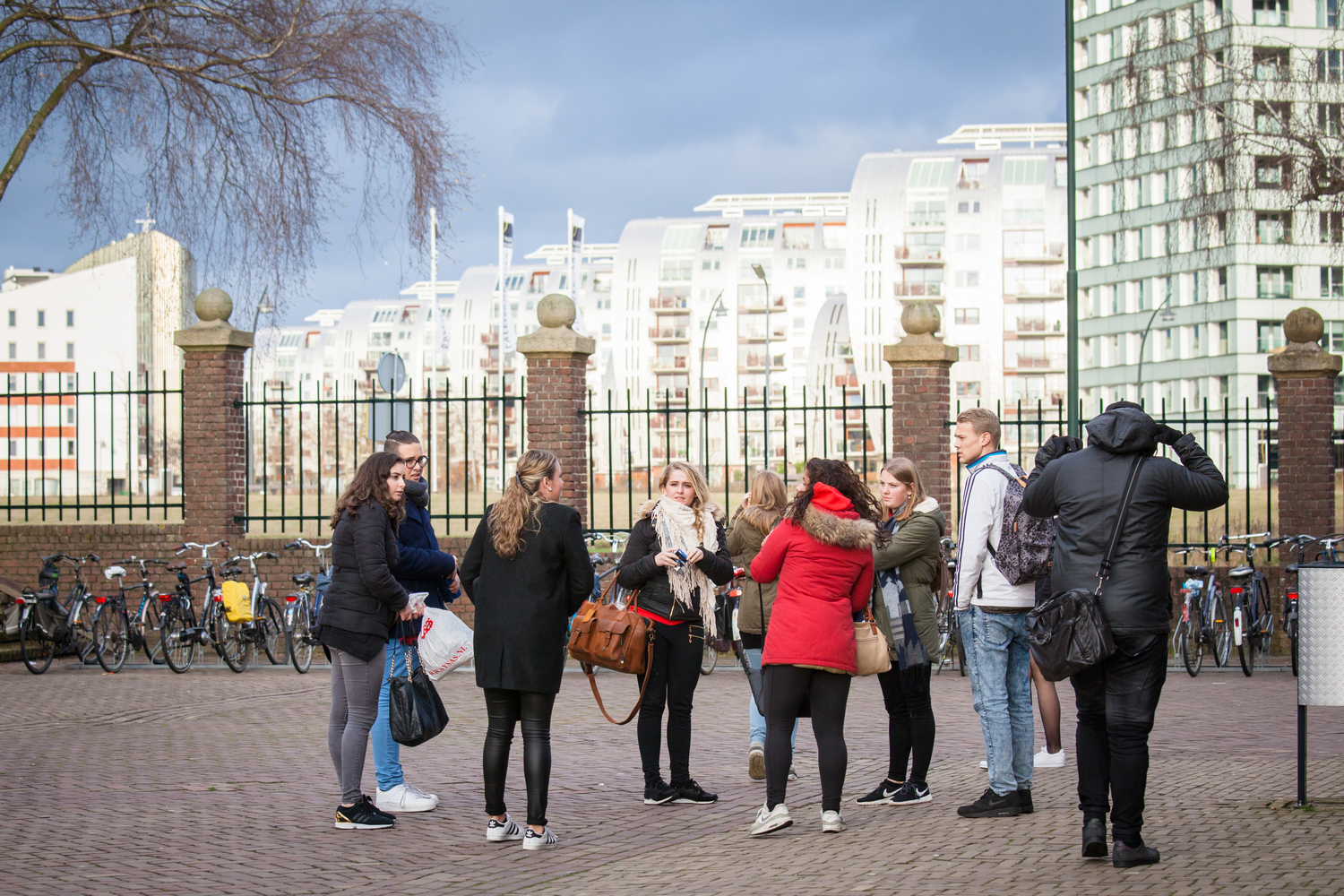 Een groep mensen die deelnemen aan VO-gerelateerde buitenschoolse activiteiten, staande voor een bakstenen gebouw. School en Veiligheid