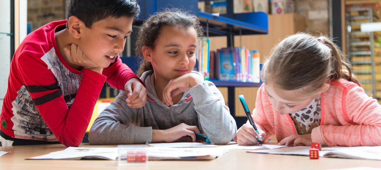 Drie kinderen zitten aan een tafel in een bibliotheek, bezig met een project over gezonde relaties en seksualiteit op school. School en Veiligheid