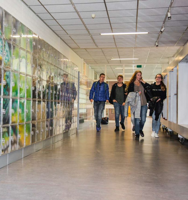 Een groep mensen, bestaande uit studenten en personeelsleden, loopt door een helder verlichte gang van een school. De school stelt haar leerlingen in staat verantwoordelijke en zorgzame individuen te worden door de sociale veiligheid te bevorderen School en Veiligheid