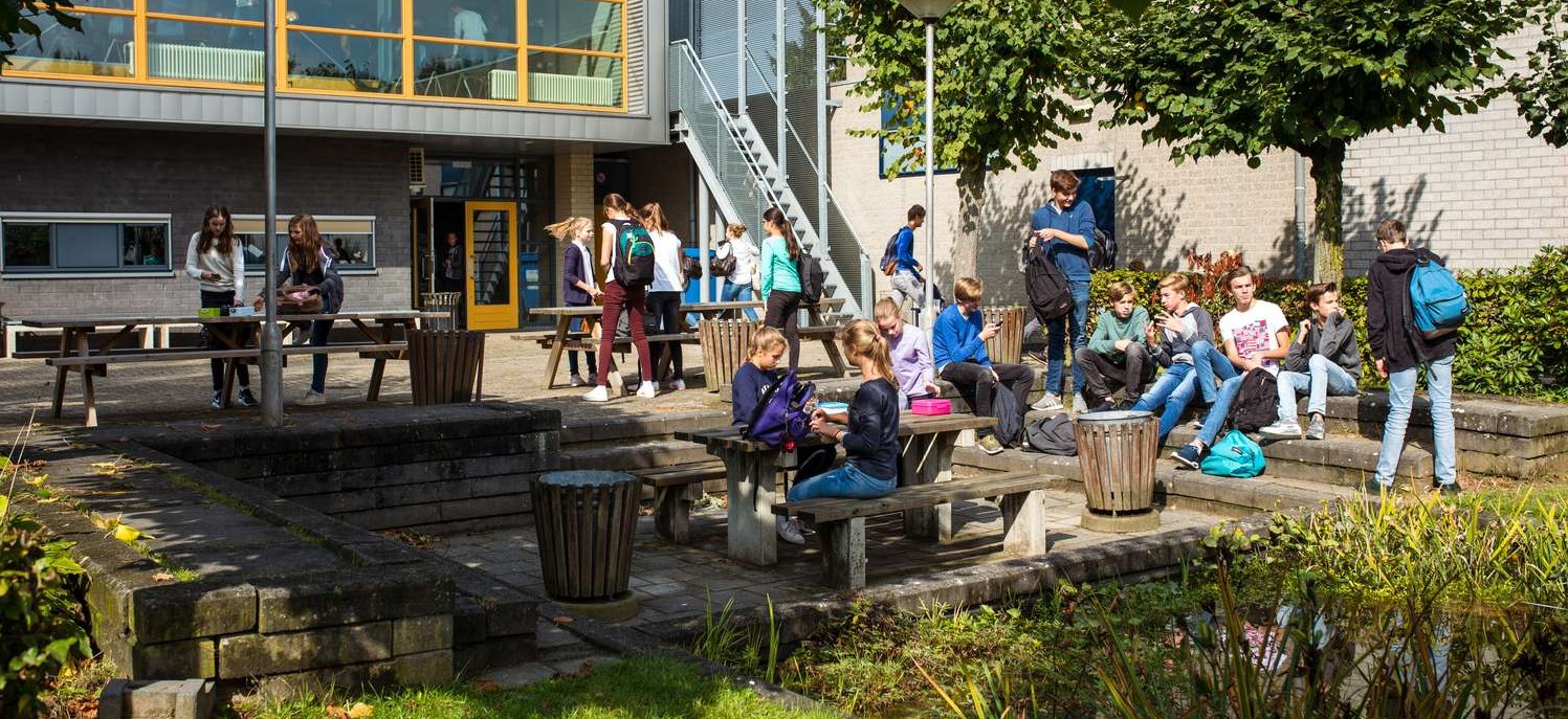 Studenten zitten en staan buiten een schoolgebouw, sommigen lezen en anderen praten over de nieuwste uitdagingen op sociale media. De setting bestaat uit banken, tafels en weelderig groen. School en Veiligheid