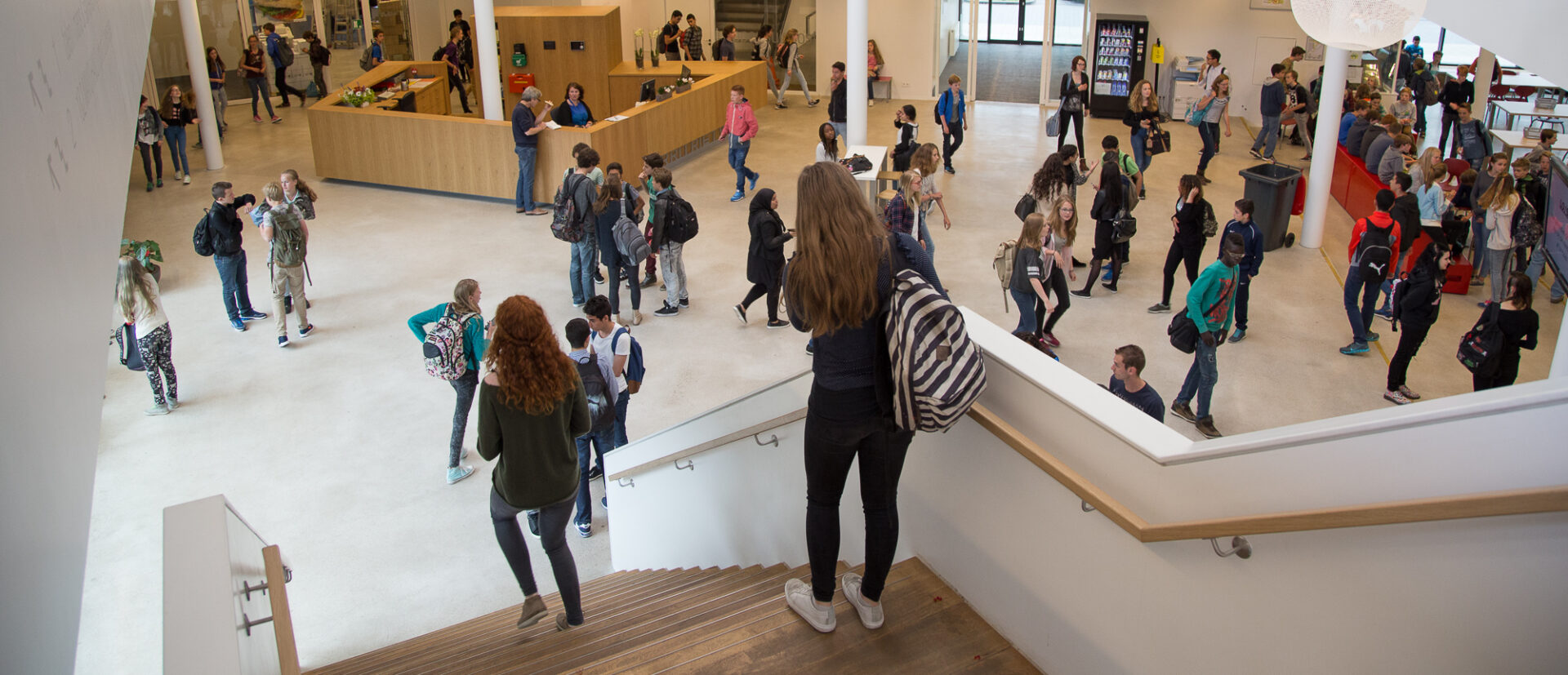 Een ruim schoolatrium met groepen studenten die staan, lopen en gezellig samenzijn. Een trap op de voorgrond daalt af in het drukke gebied, terwijl prominent een spandoek hangt ter promotie van een veiligheidswebinar over veiligheid op school. School en Veiligheid